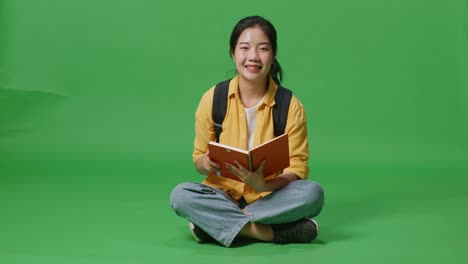 full body of asian woman student with a backpack reading a book then smiling to camera while sitting in the green screen background studio