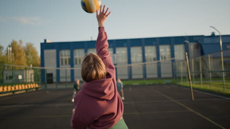 back view of volleyball player slamming the volleyball on outdoor court, with blurred people in the distance and a building in the background