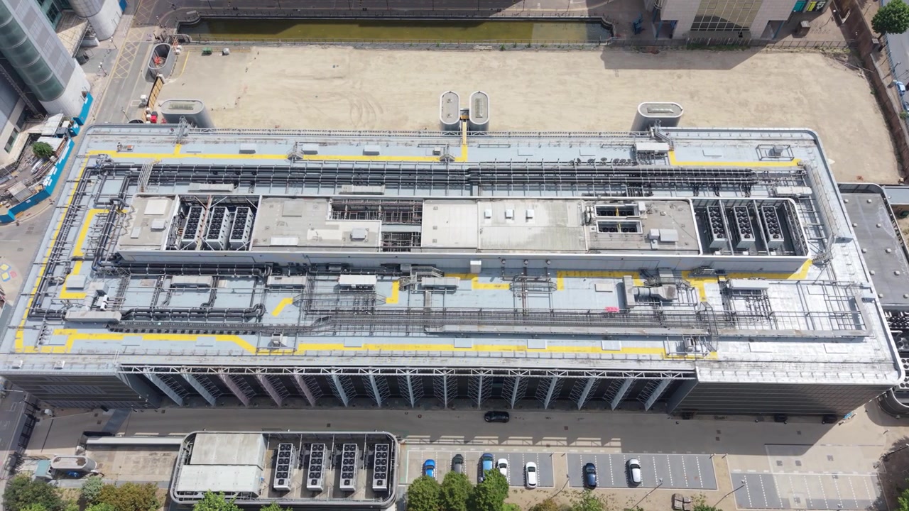 Et luftfoto viser et stort datacenteranlæg med omfattende udstyr på taget og tilstødende parkeringspladser. Trafik bevæger sig på en nærliggende vej