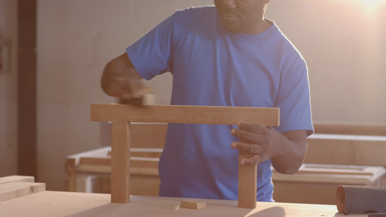 Working African American male coworkers sanding wood plank in workshop, with sanding block, goggles
