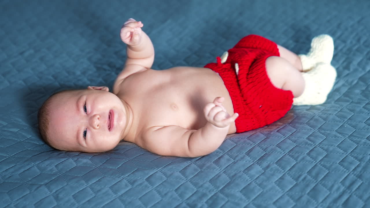 Happy little baby in red shorts with white buttons beats his feet and hands. Active kid in costume at grey backdrop.