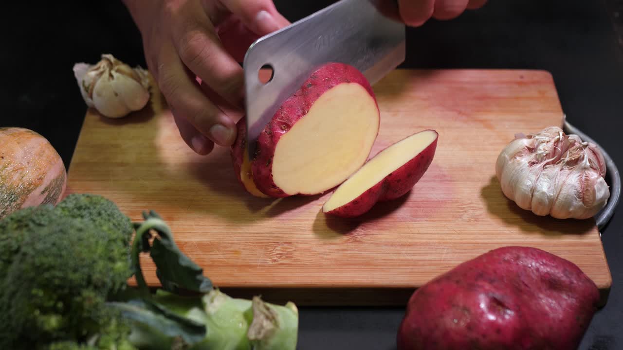 Close-up shot of hands slicing a red potato on a wooden cutting board, surrounded by fresh garlic and broccoli. The detailed view highlights crisp textures under bright lighting