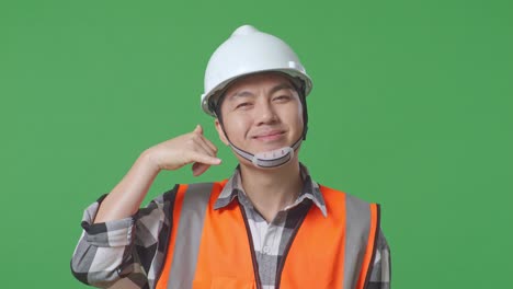 close up of asian male engineer with safety helmet smiling to camera and making call me gesture while standing in the green screen background studio