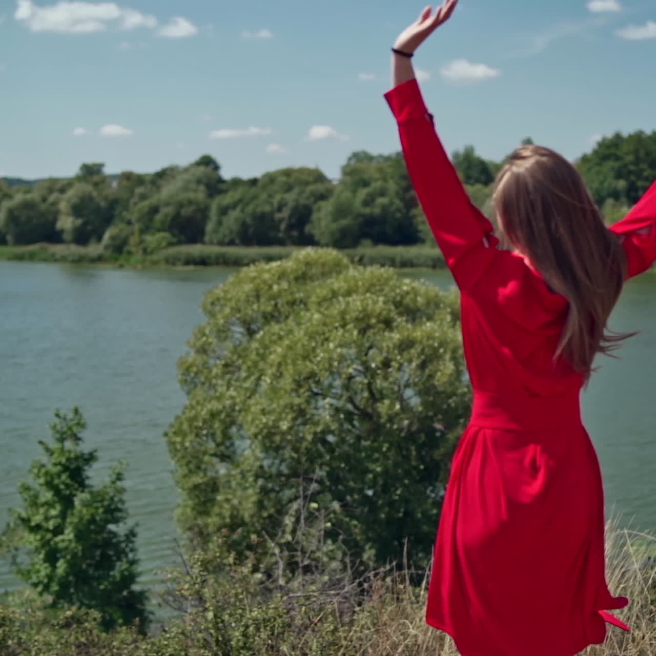 Happy woman enjoying nature in summer. Beautiful girl in red dress running and jumping merrily on the blue river background. Summer vacation.