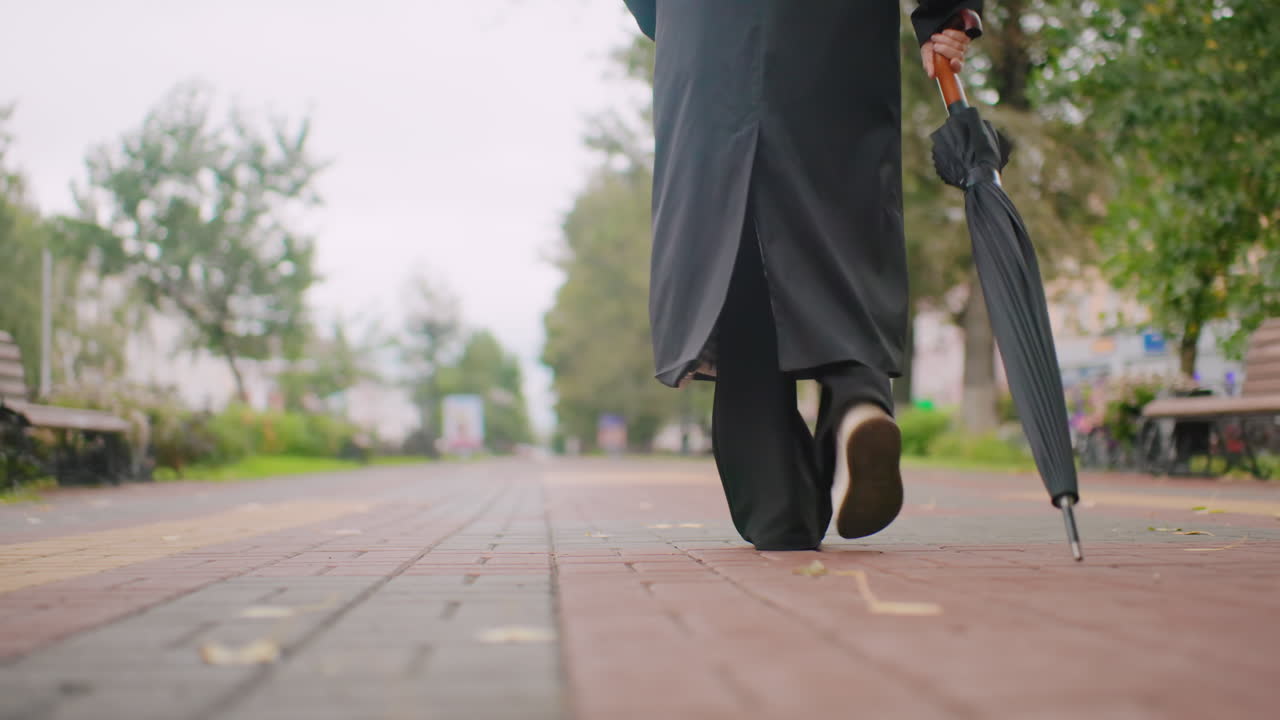 Woman dressed in black coat holding closed umbrella walks along paved pathway during cloudy autumn day, close-up of lower body and legs with atmospheric city park background highlighting seasonal solitude