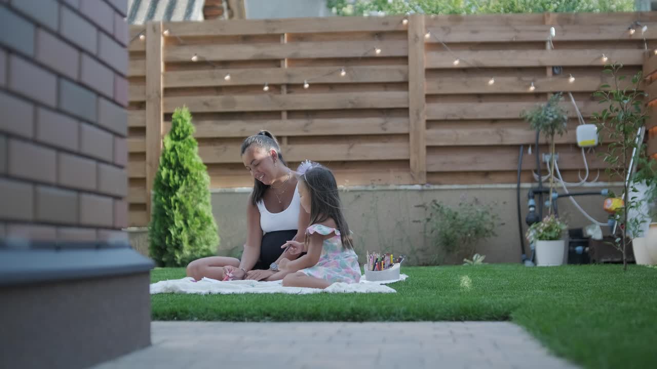 A mother and her young daughter sit on a blanket in the backyard, engaging in a drawing activity together. The outdoor scene captures a peaceful family moment, fostering creativity and bonding.