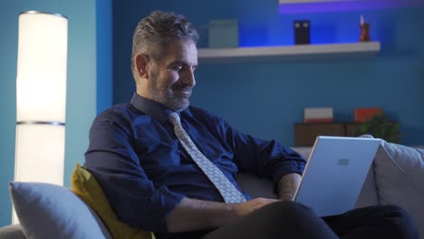 Mature-businessman-relaxing-on-sofa-working-on-laptop,-taking-notes-in-his-diary.