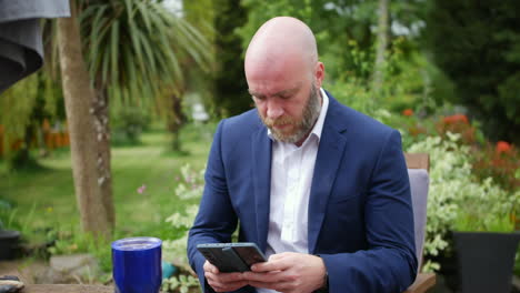 A-man-using-a-flip-phone-whilst-working-from-home-in-is-garden-with-a-coffee-cup-on-the-table-surrounded-by-greenery