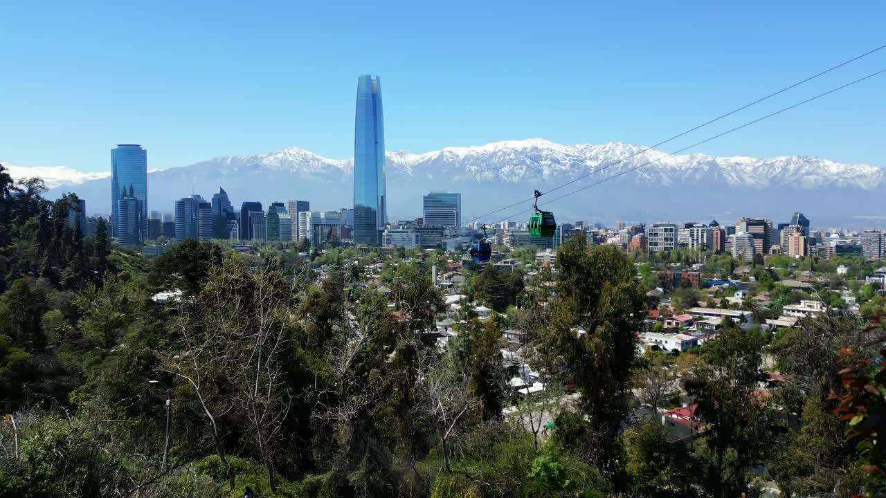 Drone aerial still of Costanera Tower and downtown Santiago with an aerial gondola cabin in the foreground and the snowcapped Andes stretching across the background under clear daytime skies