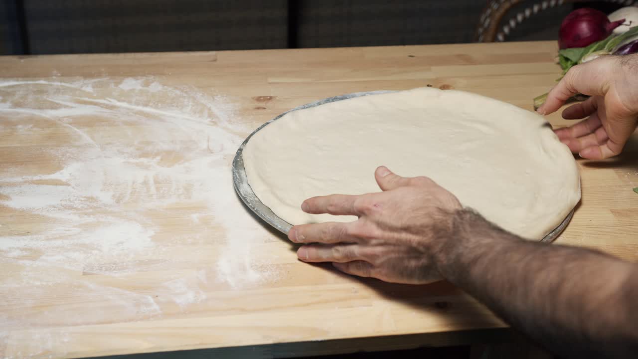 Una ripresa dall'alto delle mani di uno chef esperto che mettono l'impasto della pizza su una teglia d'acciaio su un bancone di legno, stendendolo e preparandolo per il forno.