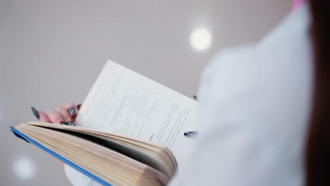 rear partial view of woman reading book while seated on bench, light reflection on tiles creates a soft ambiance, black polished nails contrast with crisp pages