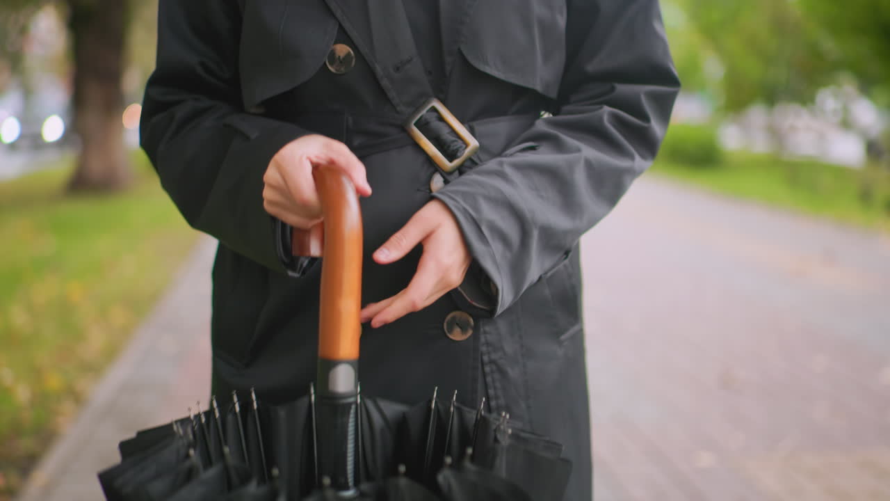 Close up of person in black coat holding closed black umbrella with wooden curved handle while walking on paved path in park symbolizing preparation, anticipation, weather protection, lifestyle