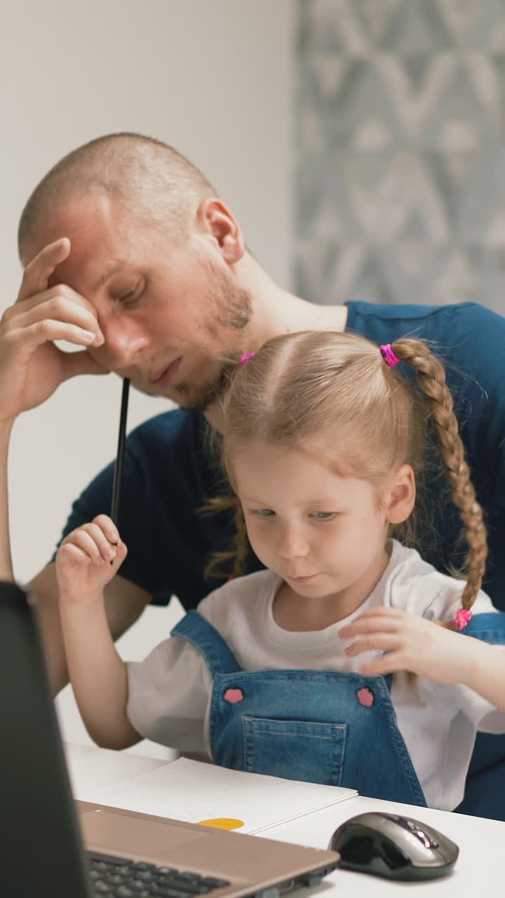 tired young father sits at table with pretty little girl drawing computer mouse near modern laptop in light room staying home at quarantine time