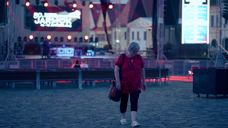 older caucasian woman wearing glasses and red t-shirt walking away from stage with flickering red and white lights and smiling in slow motion in an old historical city bardejov in slovakia