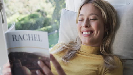 Happy-biracial-woman-sitting-next-to-window-reading-book-at-home,-slow-motion