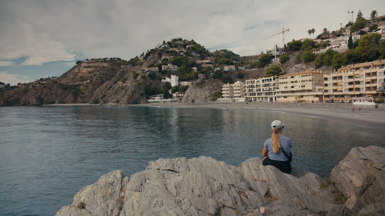 Wide establishing shot of a woman sitting on rocks, overlooking a tranquil coastal town nestled in the hills above a calm beach