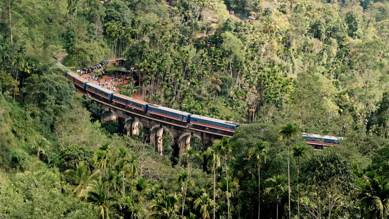 A colorful passenger train passes over the historic Nine Arch Bridge near Ella, Sri Lanka, framed by lush tropical vegetation and the green hills of the central highlands.