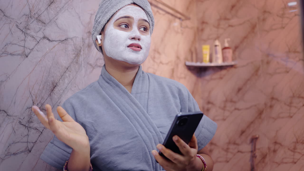 Giovane donna con maschera facciale e auricolari wireless, che parla al telefono durante una rilassante routine di cura della pelle in bagno