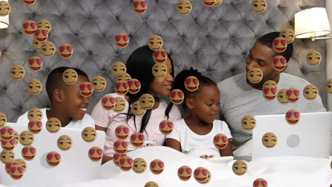 múltiples emojis de cara flotando contra la familia usando dispositivos electrónicos en la cama