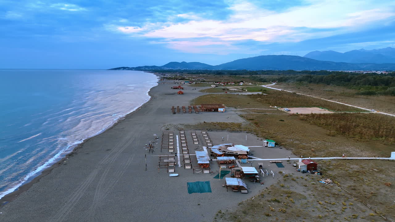 Wide line of the sandy beach at the coast of the Adriatic Sea. Approaching the tourist infrastructure with sunbeds and bars. Mountain range at backdrop. Montenegro. Aerial view