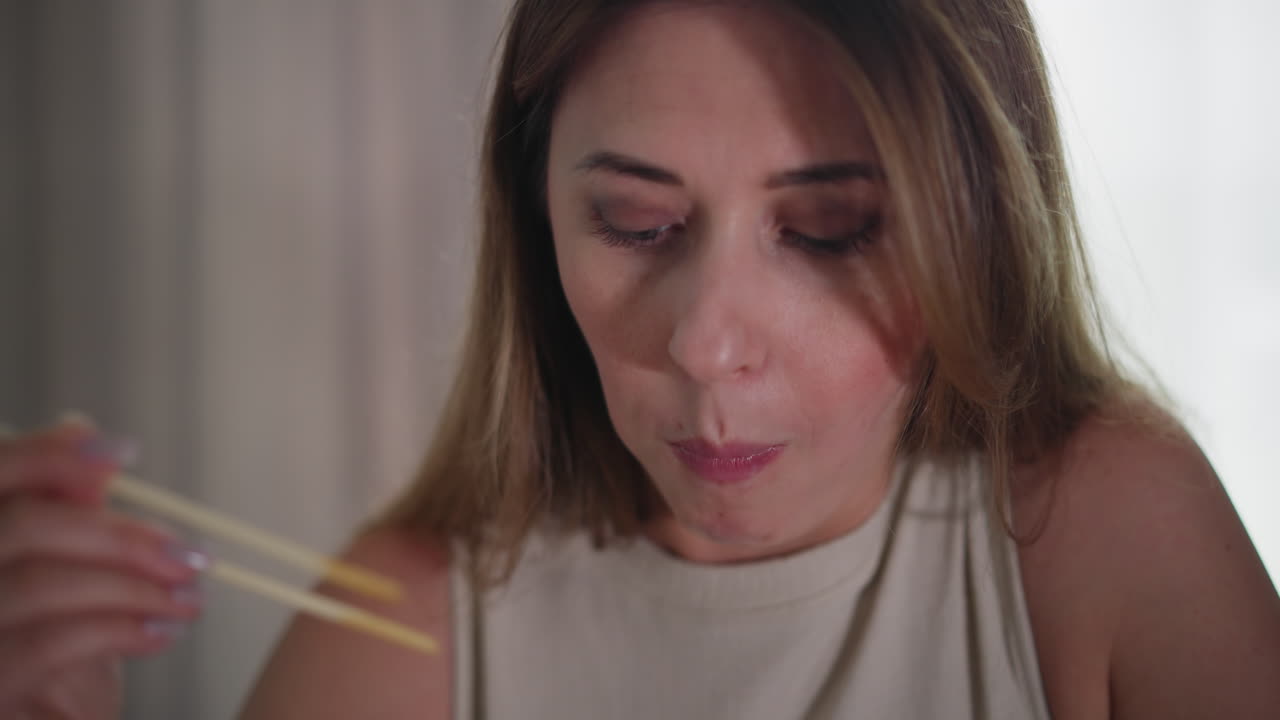 Young woman sits indoors holding meat with chopsticks, appearing thoughtful or focused while eating, natural expression, soft lighting on face, wearing sleeveless to