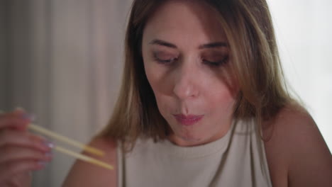 young woman sits indoors holding meat with chopsticks, appearing thoughtful or focused while eating, natural expression, soft lighting on face, wearing sleeveless to