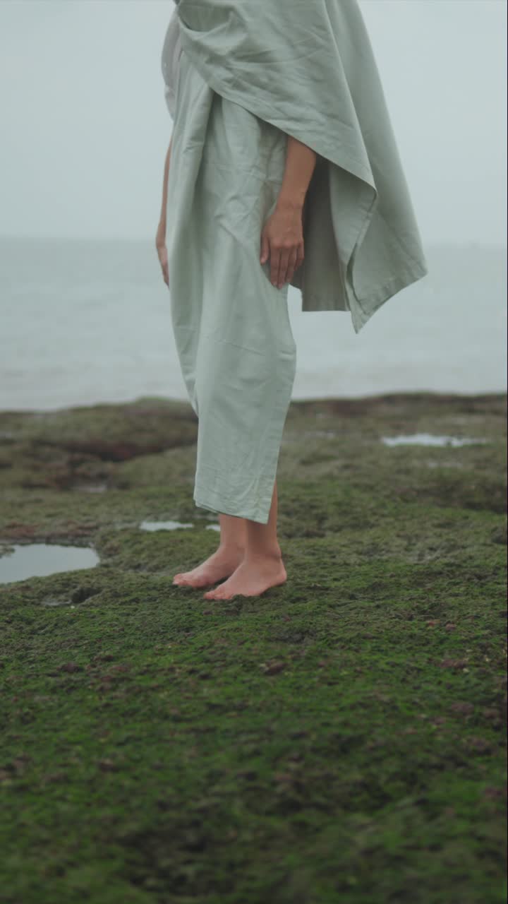 Vertical artistic shot of a woman standing barefoot on mossy grass by the sea, evoking minimalism, stillness, and contemplative coastal serenity
