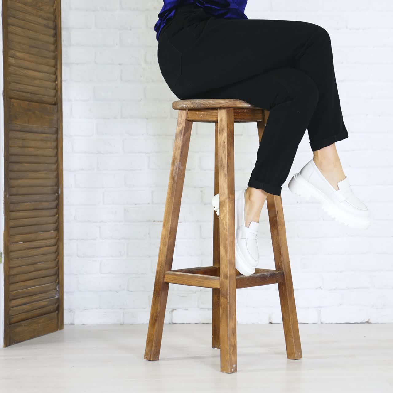 Woman in black trousers demonstrates white shoes. Lady comes and sit on the high wooden stool. White wall backdrop