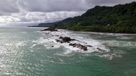 aerial orbit of turquoise sea near rocky shore and forest hillside on a cloudy day in dominicalito beach, costa rica