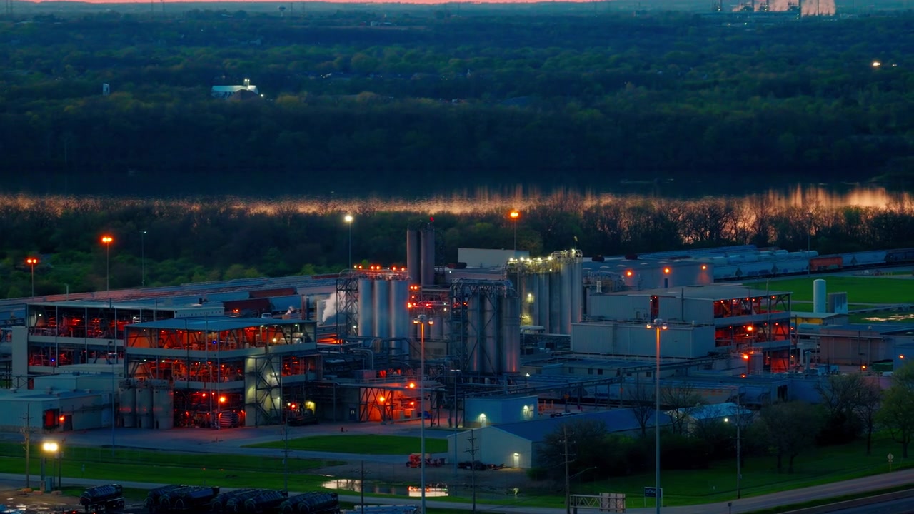 Massive, glowing chemical manufacturing facility operating at twilight, featuring complex steel infrastructure and storage silos along the river in Channahon, Illinois.