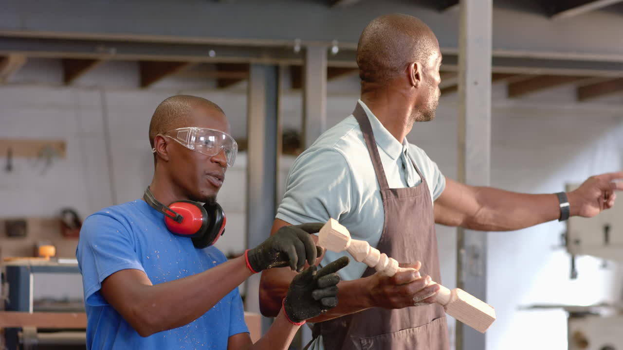 Examining African American coworkers wearing red earmuffs at shop bench, with spindle and shavings