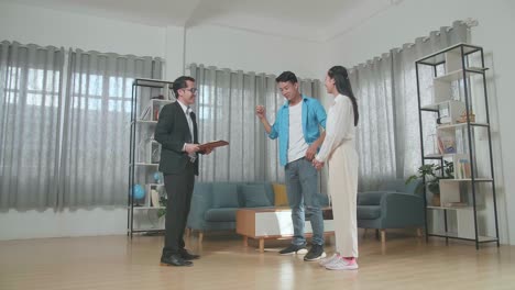 full view of asian man standing with a woman showing thumbs up gesture after receiving the keys from a real estate agent in the house for sale