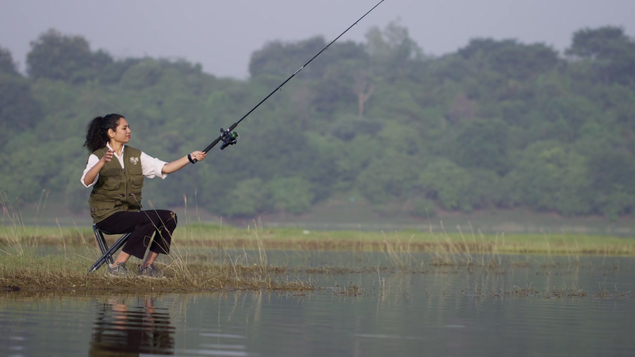 A South Asian woman fishing by a quiet lake, sitting on a chair, casting a rod, and waiting patiently in calm rural nature and daylight view calm