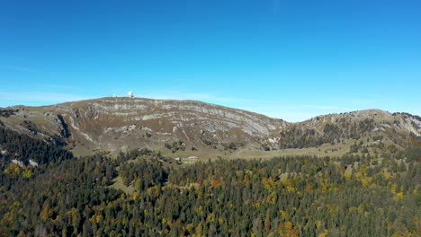 A-drone-flies-over-an-alpine-forest-with-views-across-a-large-lake-and-in-the-distant-haze-a-majestic-view-of-the-Mont-Blanc-massive-and-high-peaks-of-the-Swiss-and-French-alps