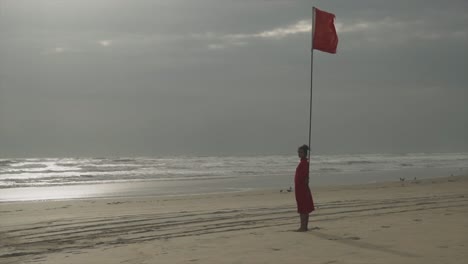 A-woman-in-a-red-dress-stands-on-an-empty-beach-during-sunset,-leaning-against-a-long-mast-with-a-red-flag