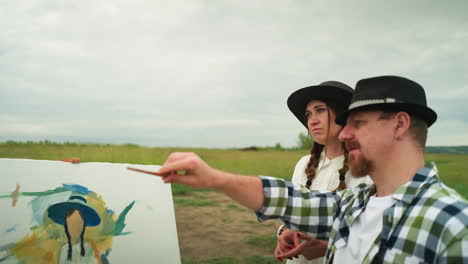 a craftsman in a checkered shirt and hat points with a brush, explaining something to a woman beside him, in a white dress and hat. they are standing in a grassy field, with a canvas in the background