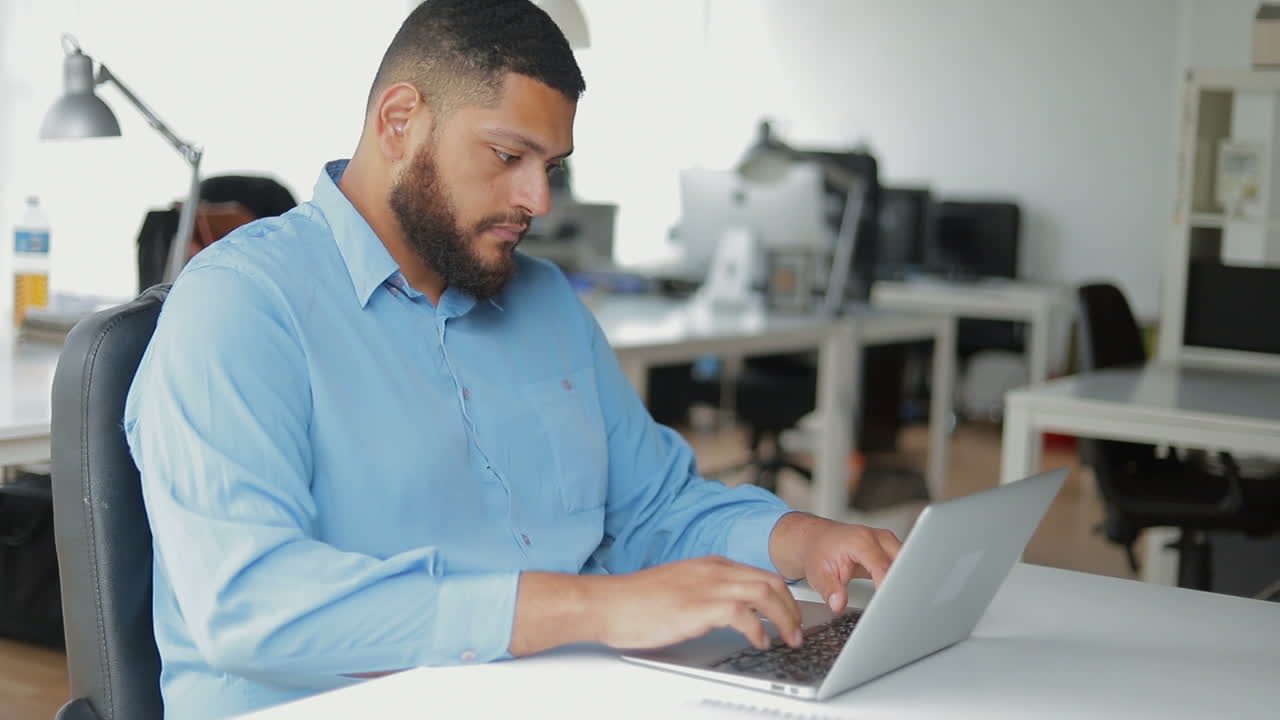 un joven barbudo pensativo escribiendo en una computadora portátil.