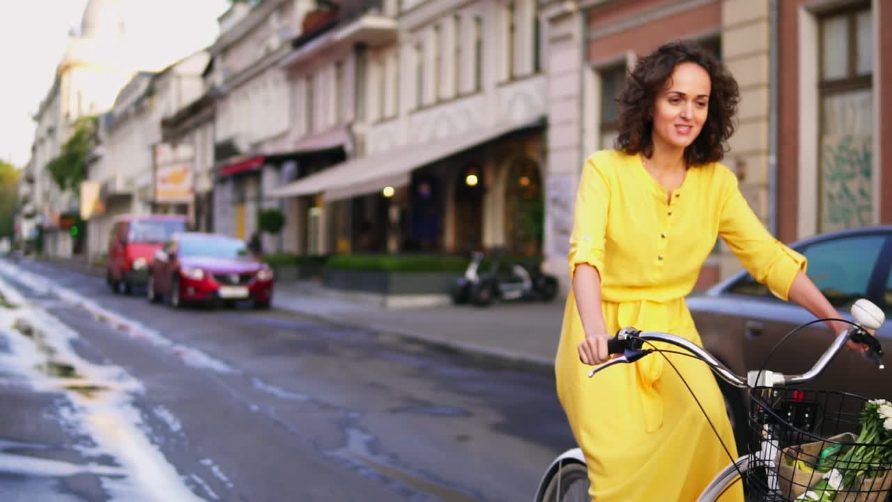Beautiful woman riding a city bicycle with a basket and flowers in the city center while talking to someone. Steadicam shot