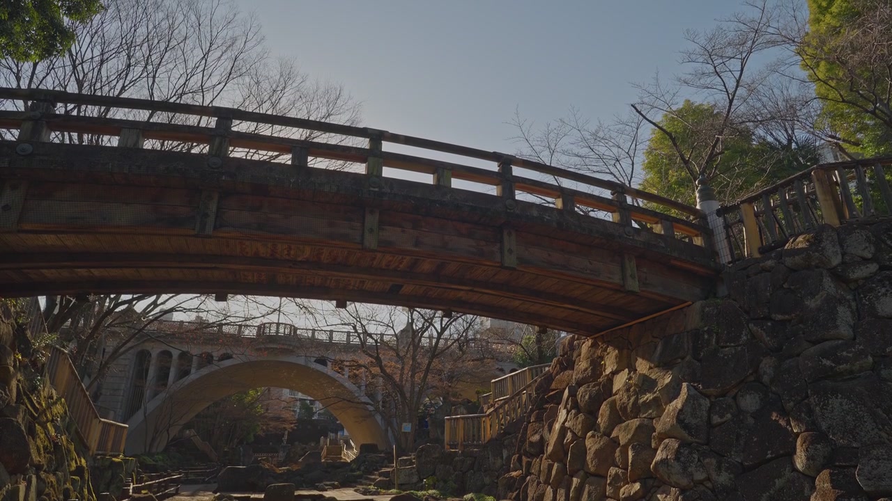 L'arco in legno del Ponte Funakushi incornicia un secondo ponte ad arco sullo sfondo, sopra un terreno roccioso del giardino con muri in pietra e alberi spogli, catturato come un'inquadratura grandangolare statica dal basso in una calda luce diurna