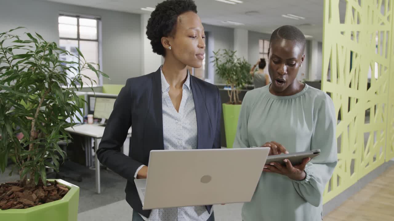 Due donne d'affari afroamericane discutono di lavoro, utilizzando un laptop e un tablet in ufficio al rallentatore.
