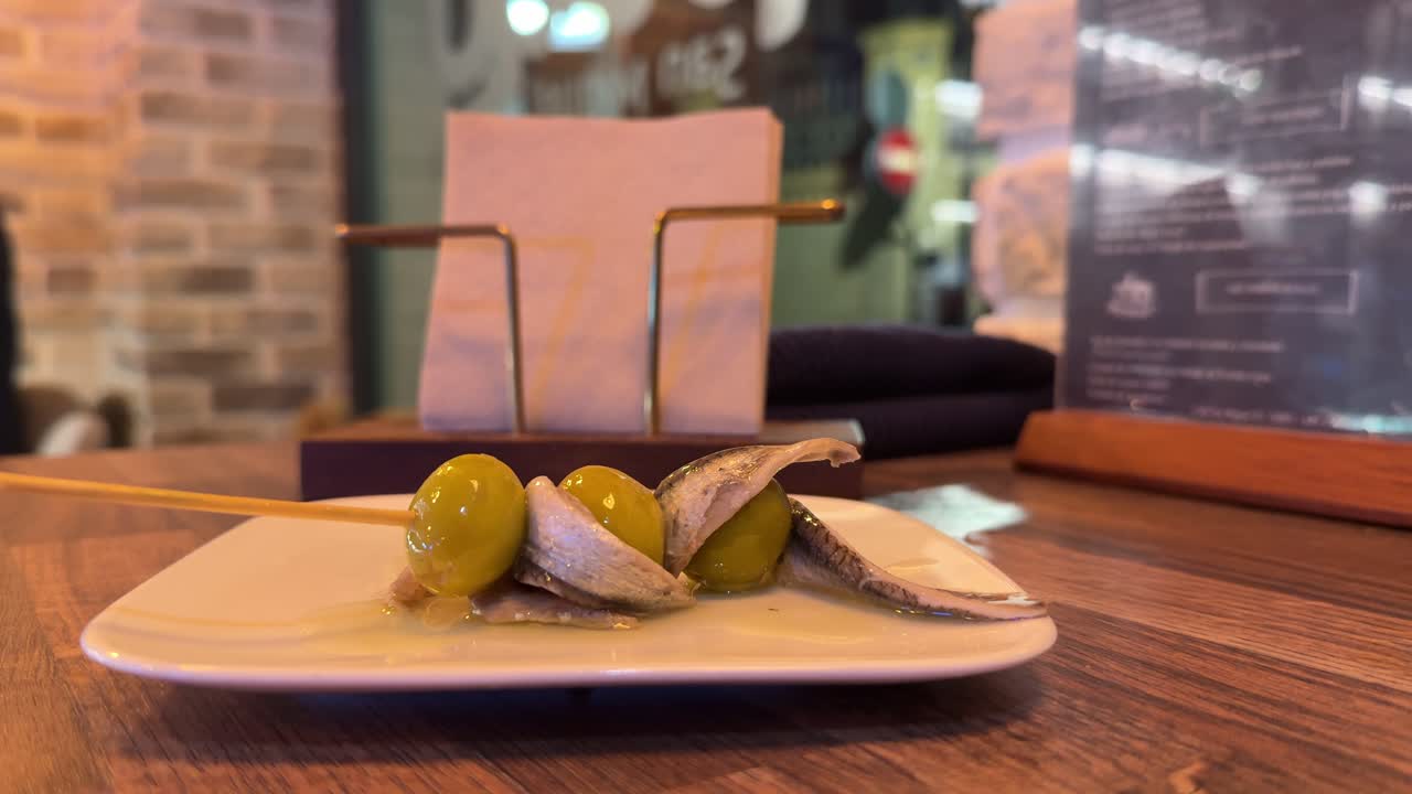 Primo piano della mano di un uomo caucasico che prende un tradizionale pincho spagnolo di acciughe e olive e una birra in un ambiente rilassato all'interno di un bar tradizionale