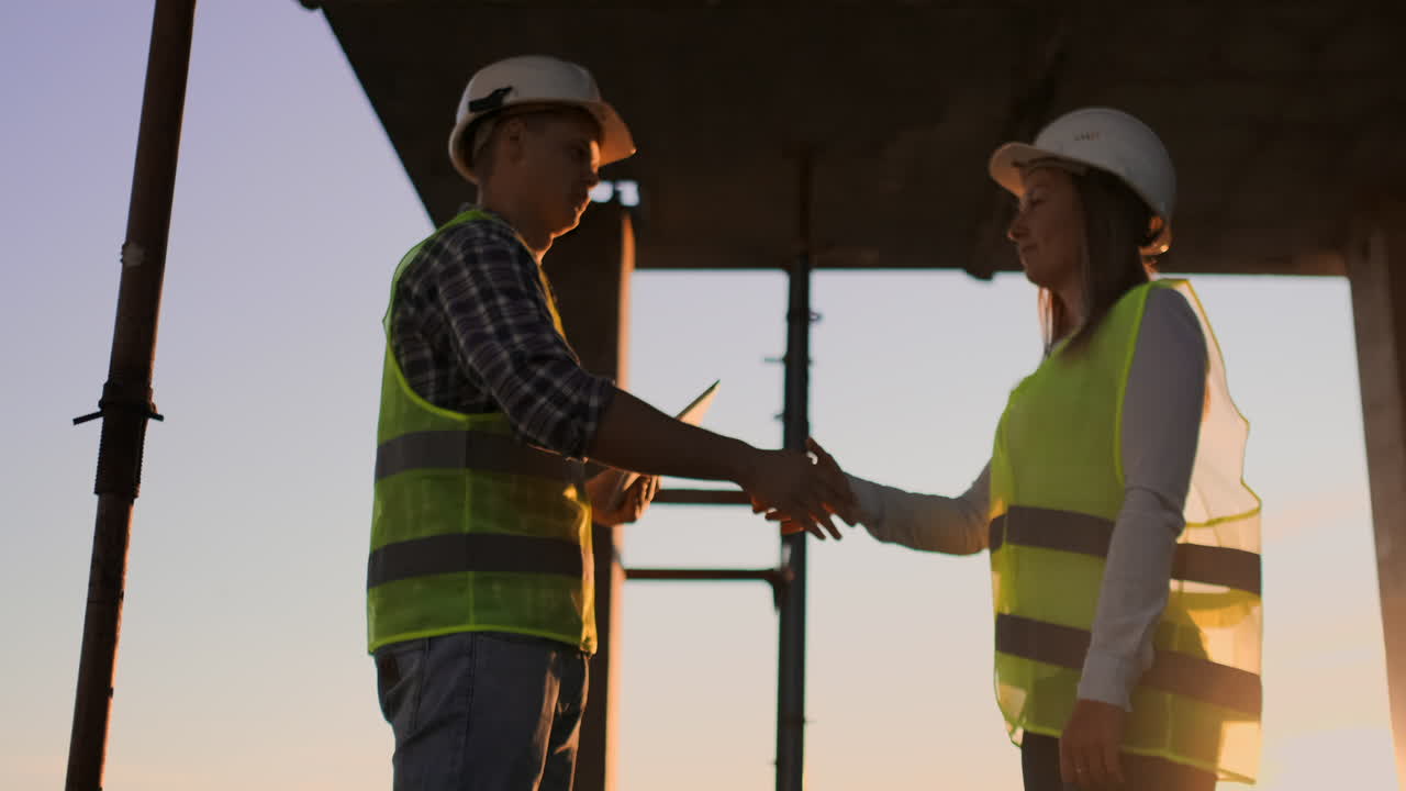 Builders man with a tablet and a woman in white helmets shake hands at sunset standing on the roof of the building