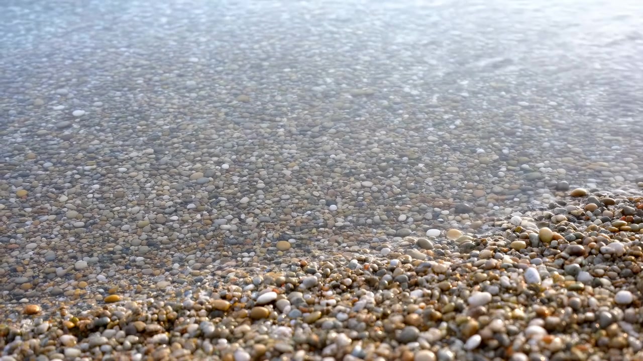 Nahaufnahme eines Küstenpanoramas mit glatten Kieselsteinen und seichtem, klarem Wasser, während eine dünne Welle über den Strand rollt und reflektierende nasse Streifen, feine Wellen und eine ruhige Meeresoberfläche hinterlässt.