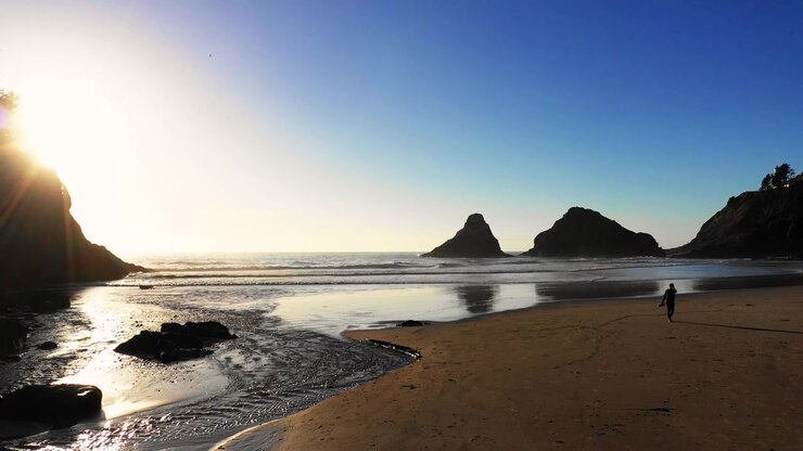 Um homem carregando uma câmera na praia, caminhando em direção à costa do Oregon durante o pôr do sol, com o céu azul e ondas calmas do oceano ao fundo.
