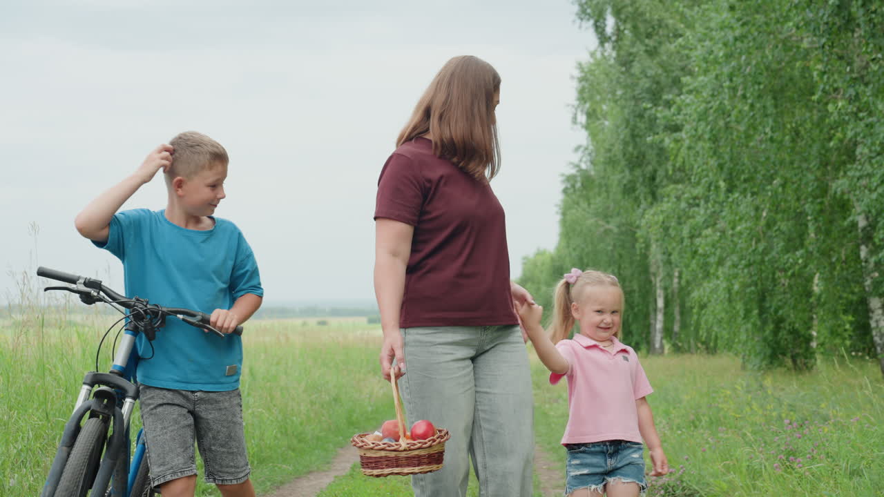자작나무가 늘어선 길을 따라 걷는 백인 엄마가 딸의 손을 잡고 있어요. 딸은 깡총깡총 뛰면서 낄낄 웃고, 십 대 아들은 자전거를 끌고 근처에 있네요. 사과 바구니가 흔들리고, 솔직한 미소가 보기 좋은 따뜻한 여름 가족의 모습이에요.