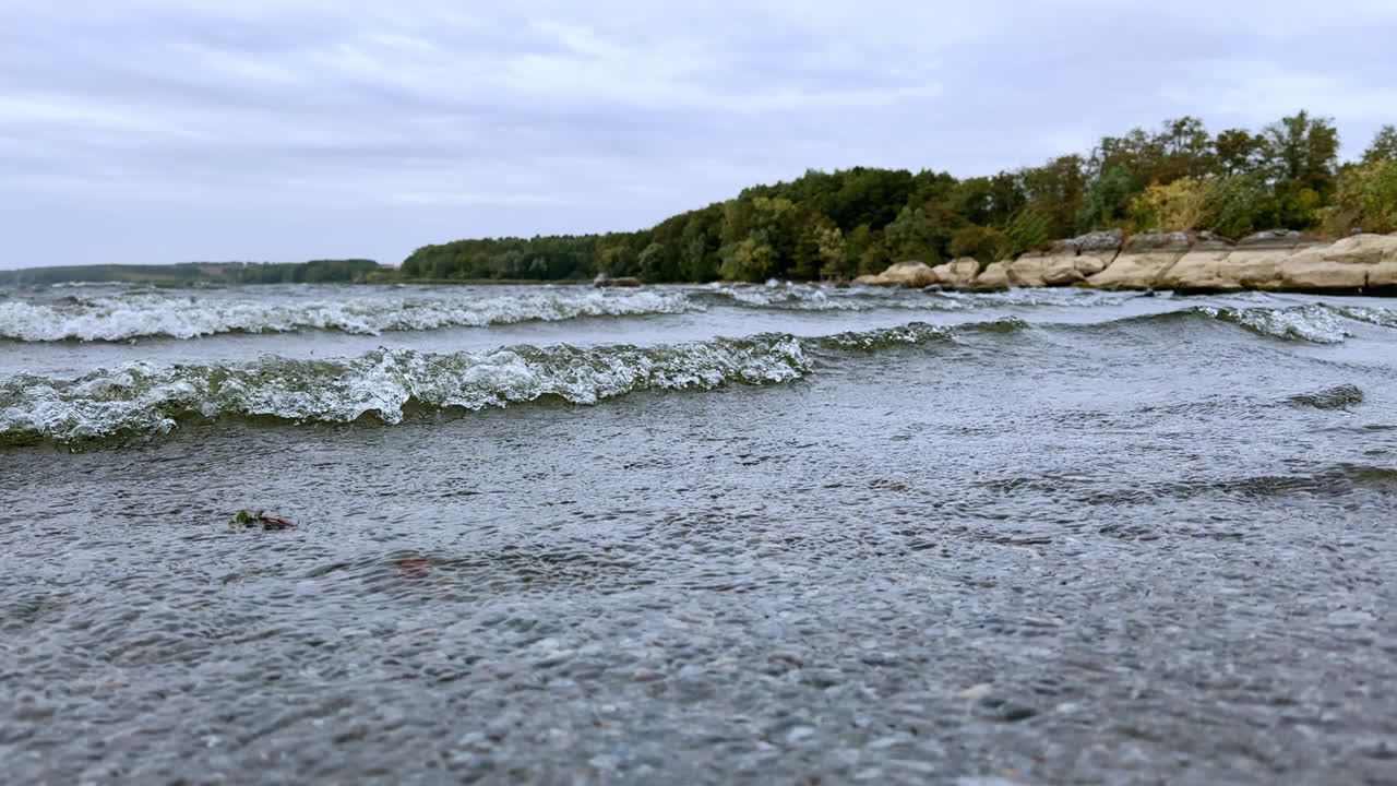 Wind sending the river waves move quickly to the shore. Close up. Rocky bank with green trees at backdrop.