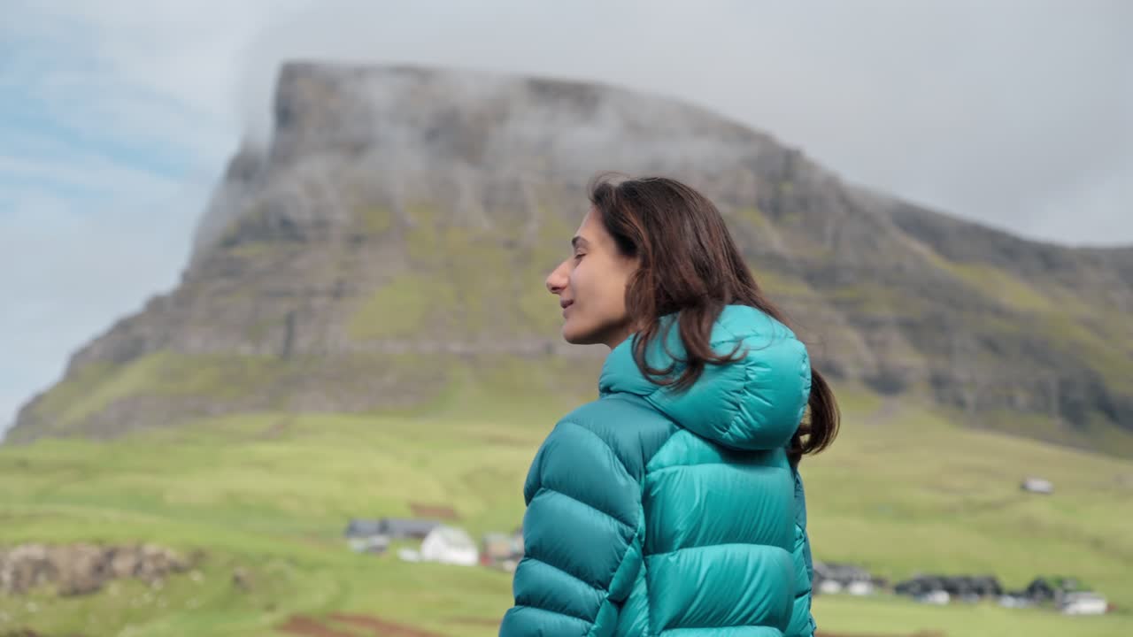 A person enjoys a serene mountain view in a green valley wearing a teal jacket outdoors