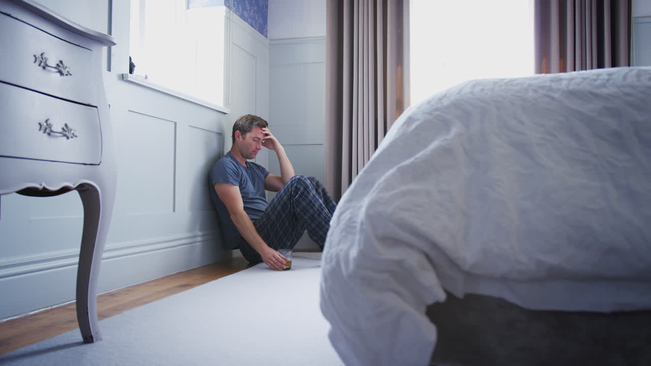 Depressed Man Wearing Pajamas Sitting On Floor Of Bedroom Holding Glass Of Whisky