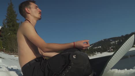 joven sin camisa respirando pesadamente, sentado en una alfombra de yoga en un paisaje nevado al lado del lago de montaña congelado se prepara para un baño de hielo