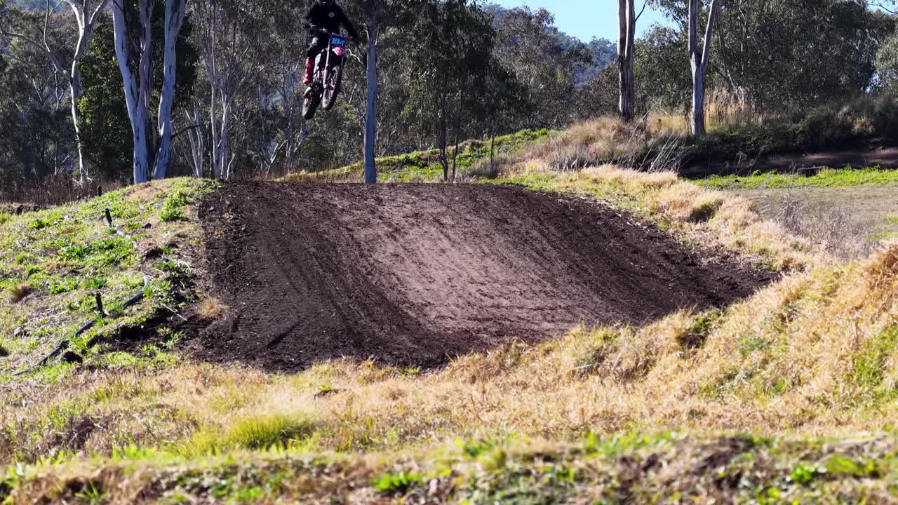 Motocross rider jumps dirt mound on sunlit outdoor track, surrounded by trees and grassy terrain